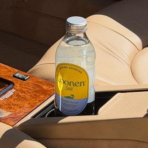 A bottle of Boonen Still sparkling water placed in the center console of a car with beige leather seats and a wood trim dashboard.