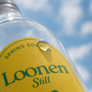 A close-up angled shot of a clear glass bottle with a yellow label reading 'Loonen Still' and 'SPRING SOURCED' in green text, set against a bright blue sky with white clouds.